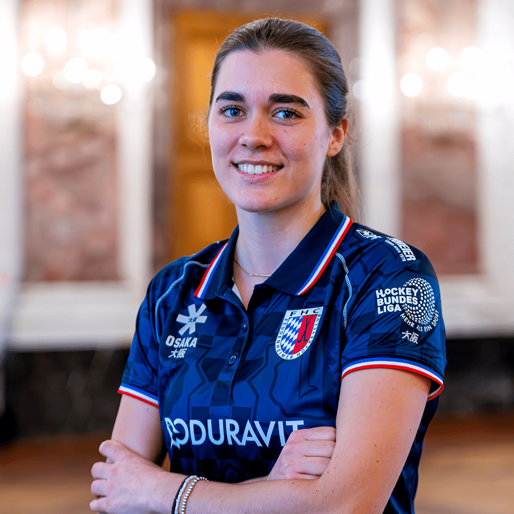 Eine lächelnde Hockeyspielerin in einem blauen Trikot des Feudenheimer Hockeyclubs posiert selbstbewusst für ein Porträtfoto in einem Saal.