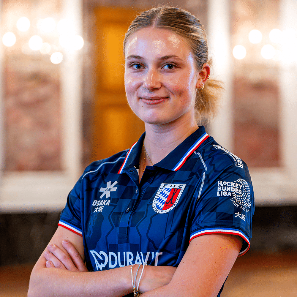 Eine junge Hockeyspielerin in einem blauen Trikot des Feudenheimer HC lächelt selbstbewusst in die Kamera, ähnlich wie beim Mannheimer Hockeyclub.