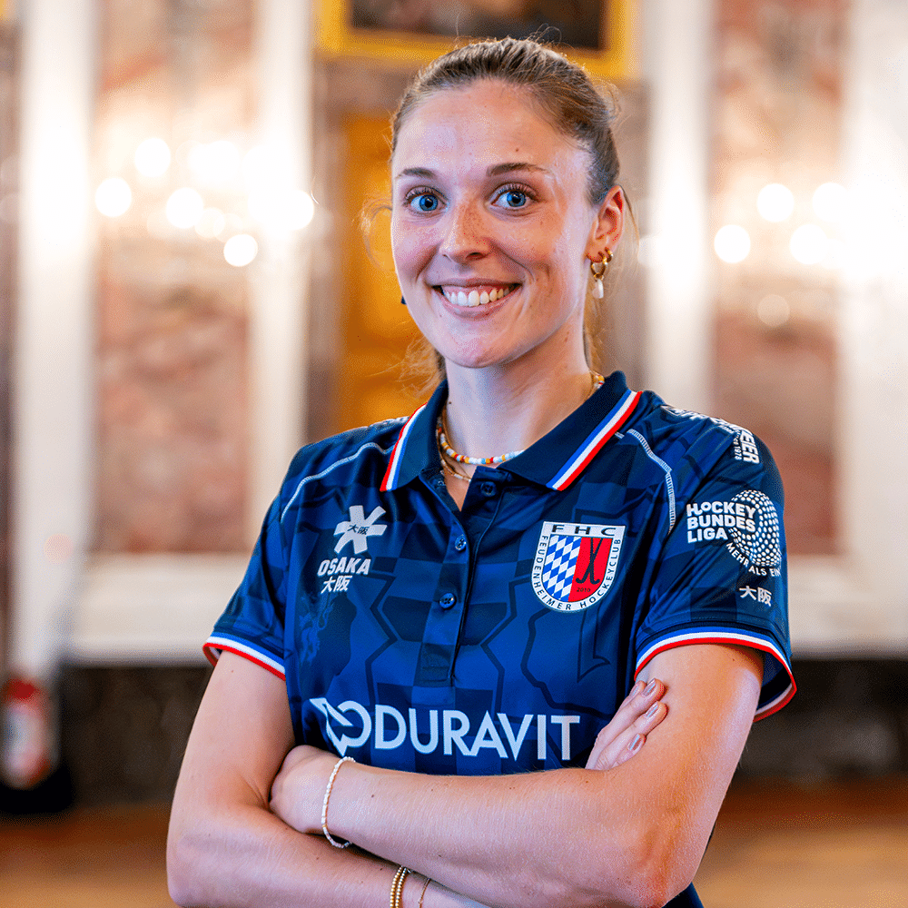 Eine lächelnde Hockeyspielerin im blauen Trikot des Feudenheimer Hockeyclubs posiert selbstbewusst für Hockey in Mannheim vor einem Saal.