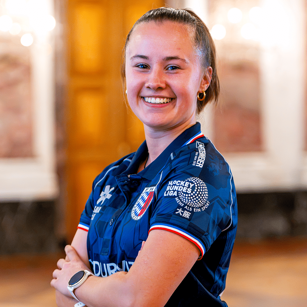 Eine lächelnde Spielerin vom MHC, dem Mannheimer Hockeyclub, trägt ihr blaues Trikot und steht für Hockey in Mannheim vor einer hellen Kulisse.