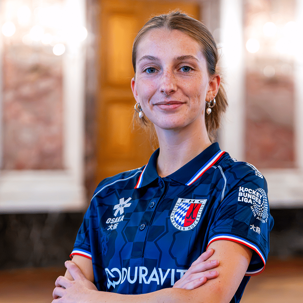 Eine junge Hockeyspielerin des Feudenheimer Hockeyclubs mit verschränkten Armen in ihrem blauen Trikot, ähnlich dem Stil beim Mannheimer Hockeyclub.