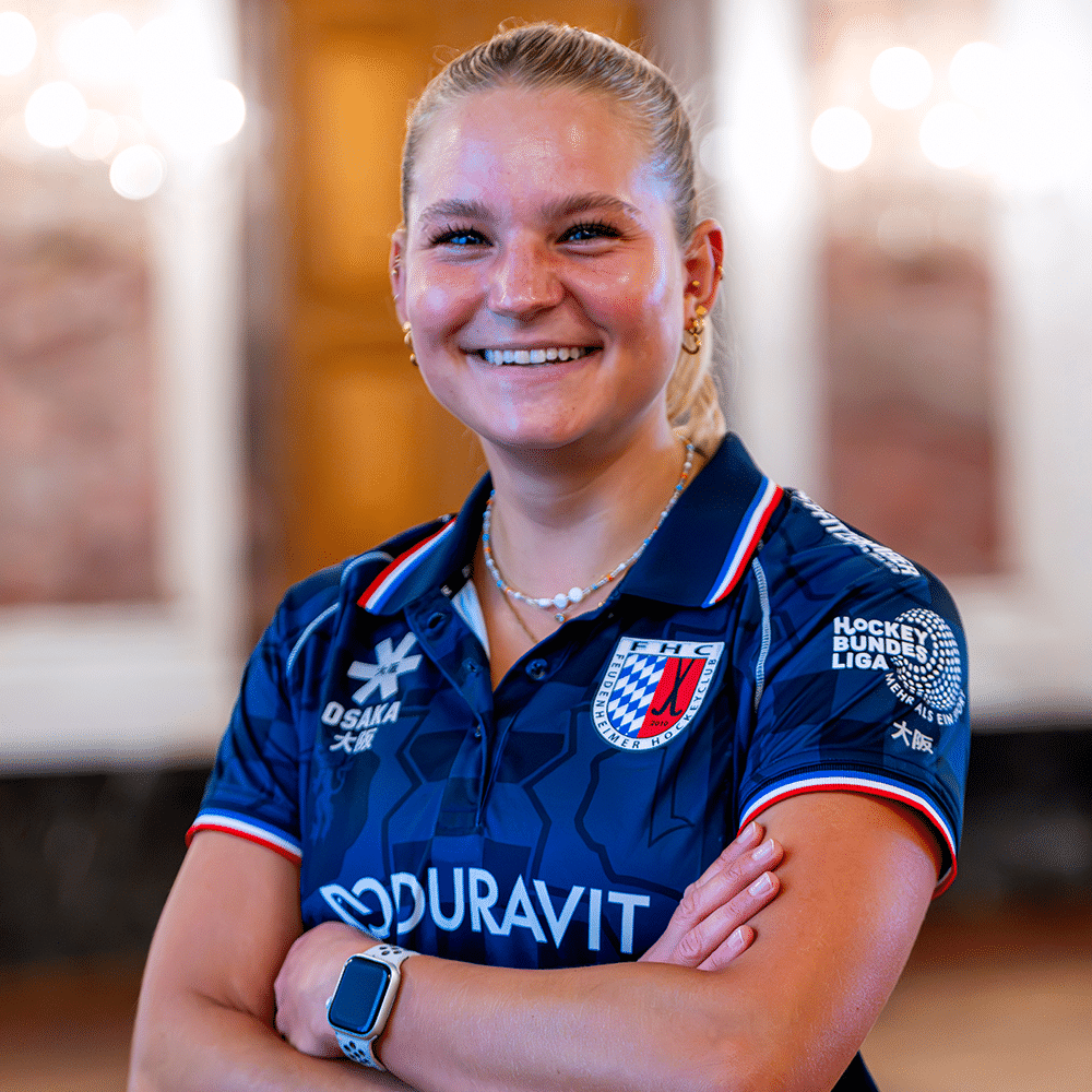 Eine lächelnde Hockeyspielerin mit blondem Pferdeschwanz trägt ein blaues Trikot des Feudenheimer Hockeyclubs und verschränkt die Arme vor der Brust.