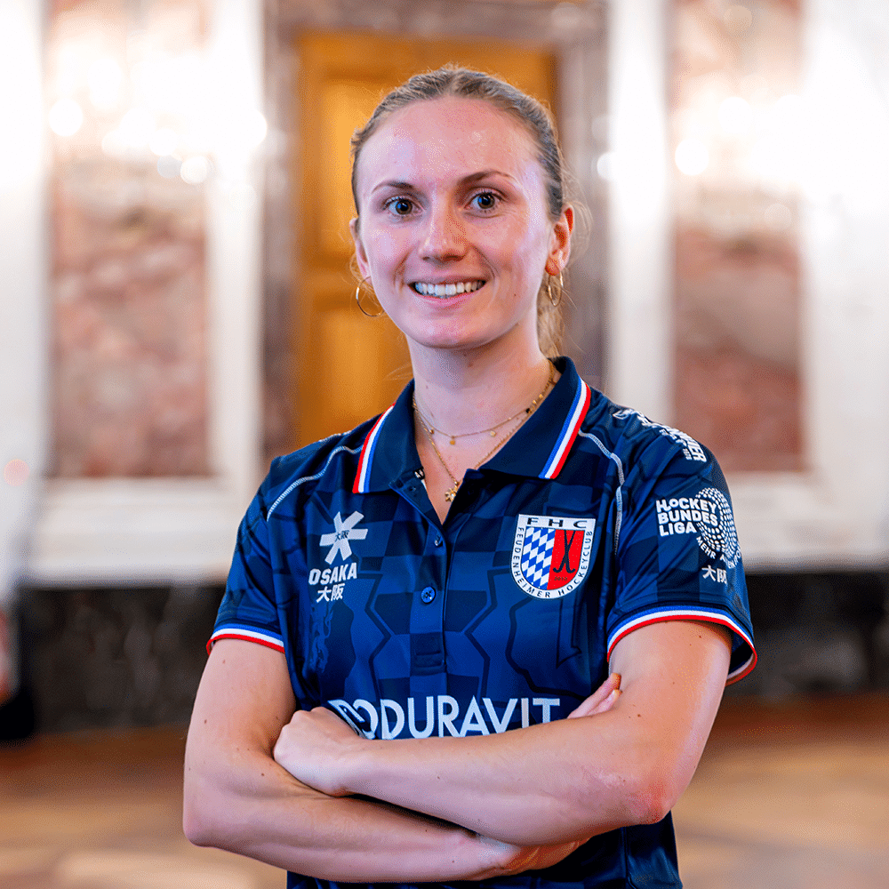 Eine lächelnde Hockeyspielerin im blauen Trikot des Mannheimer Hockeyclub vor einer eleganten Kulisse. Hockey in Mannheim wird hier repräsentiert.