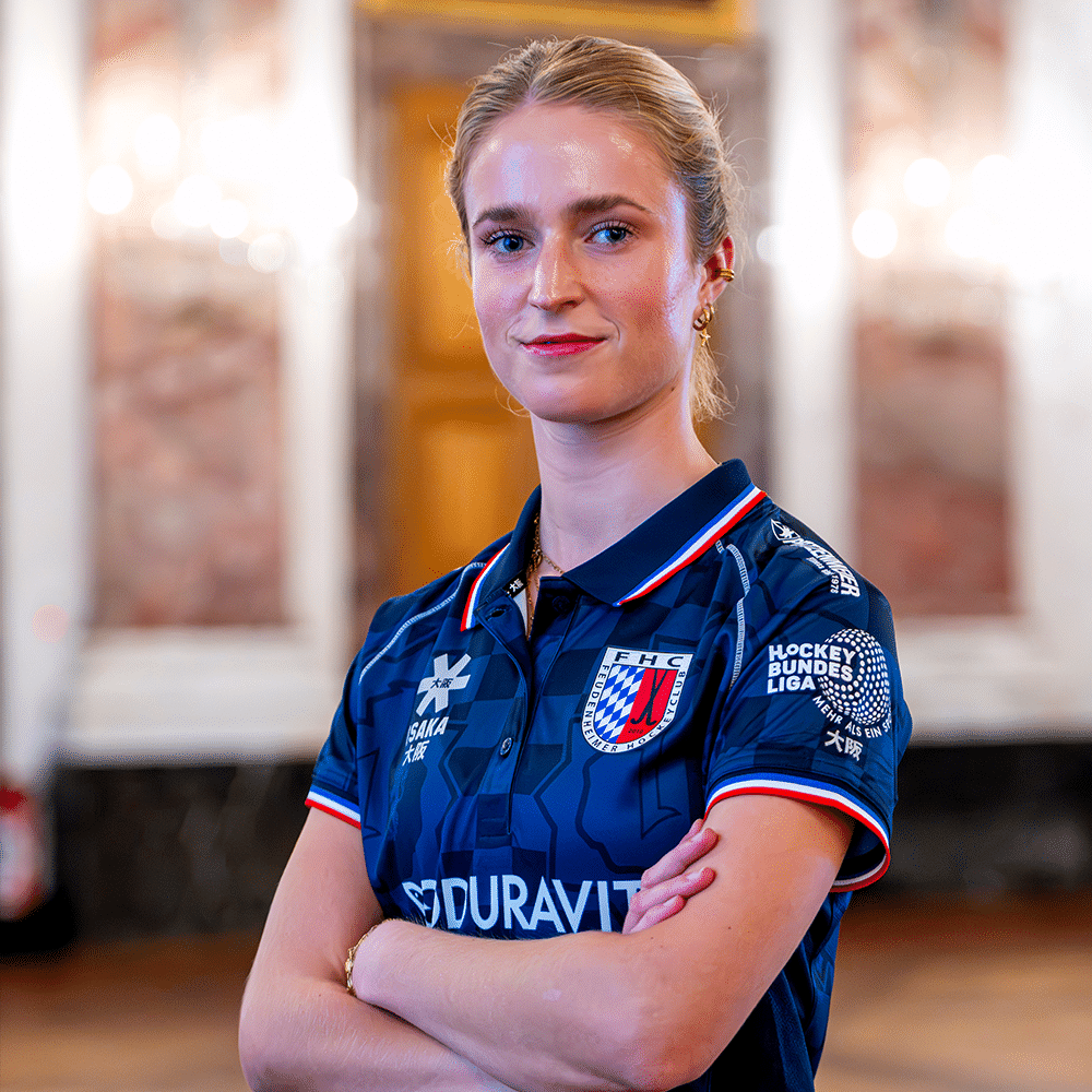 Eine junge Hockeyspielerin in einem blauen Trikot des Feudenheimer Hockeyclubs steht selbstbewusst vor einer eleganten Kulisse in Mannheim.