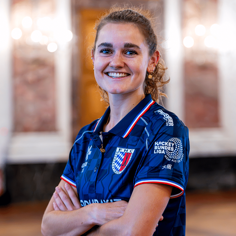 Eine lächelnde Hockeyspielerin in einem blauen Trikot des MHC steht mit verschränkten Armen vor einem eleganten, unscharfen Hintergrund.
