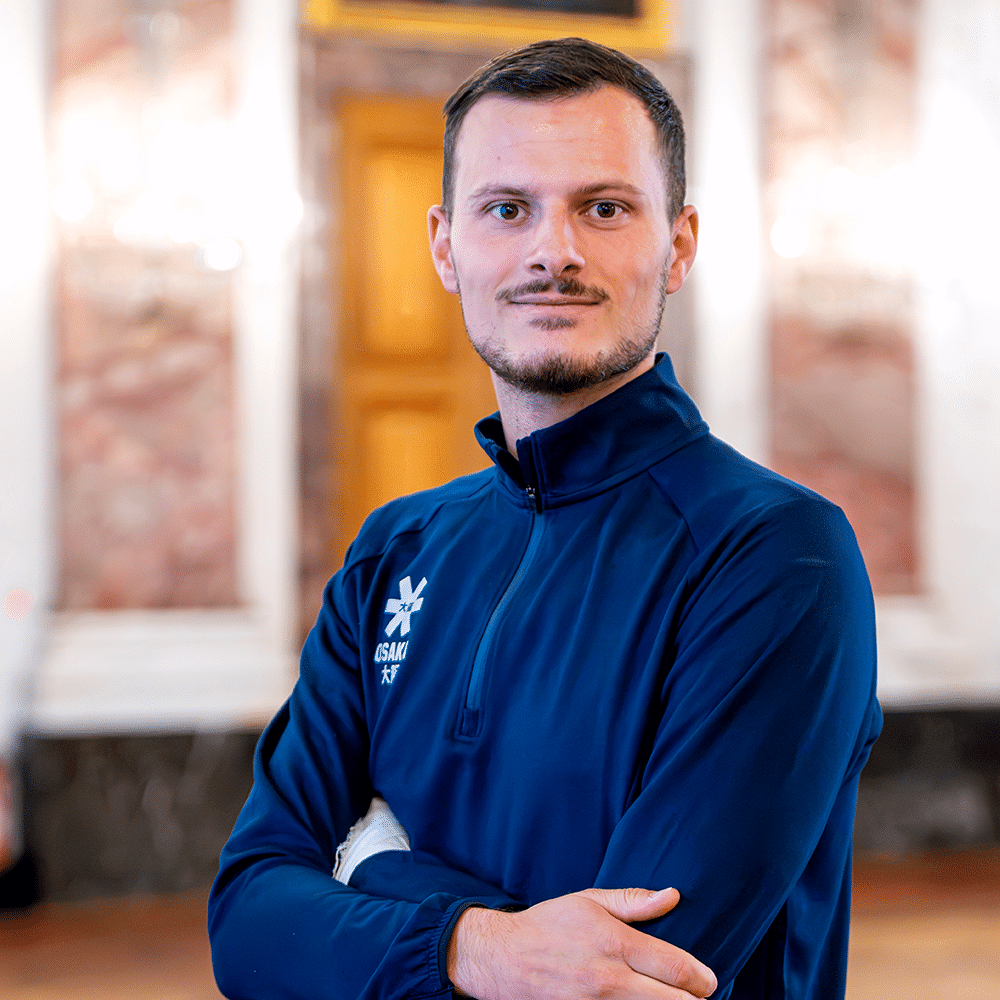 Ein junger Mann mit kurzem Haar und Bart trägt ein dunkelblaues Sportoberteil und steht vor einer klassischen Architekturkulisse in Mannheim.