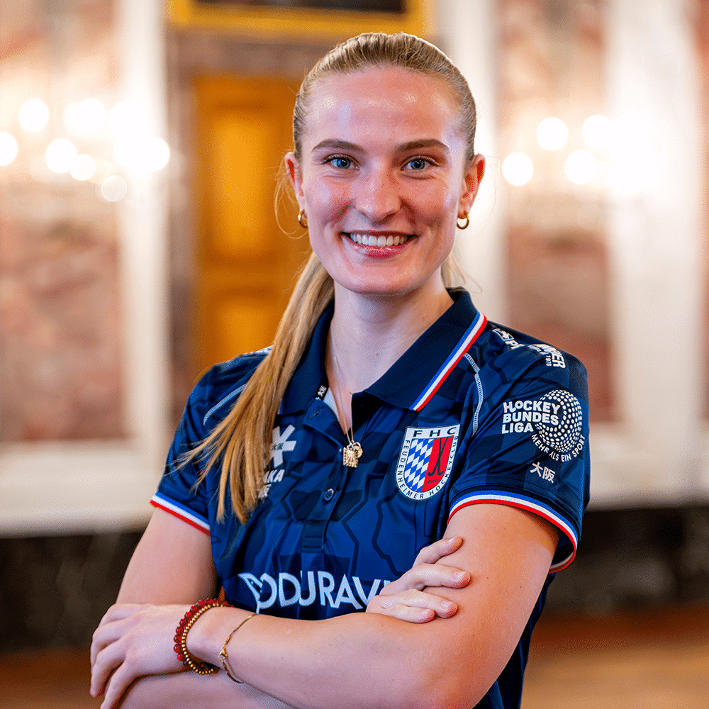 Eine lächelnde Hockeyspielerin in einem blauen Trikot des Feudenheimer Hockeyclubs steht vor einem eleganten Hintergrund und blickt in die Kamera.