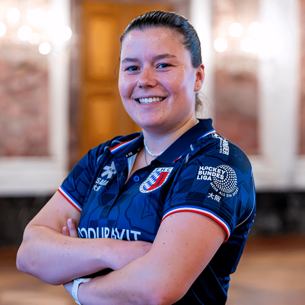 Eine lächelnde Spielerin vom MHC in ihrem blauen Vereinstrikot mit verschränkten Armen. Hockey in Mannheim wird durch den Mannheimer Hockeyclub repräsentiert.