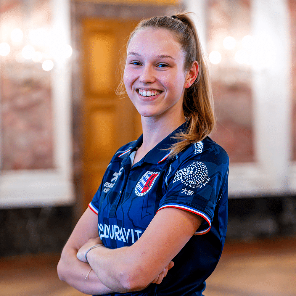 Eine lächelnde Hockeyspielerin vom Mannheimer Hockeyclub trägt ein blaues Trikot mit MHC-Logo und Sponsorenaufdrucken vor einem eleganten Saal.