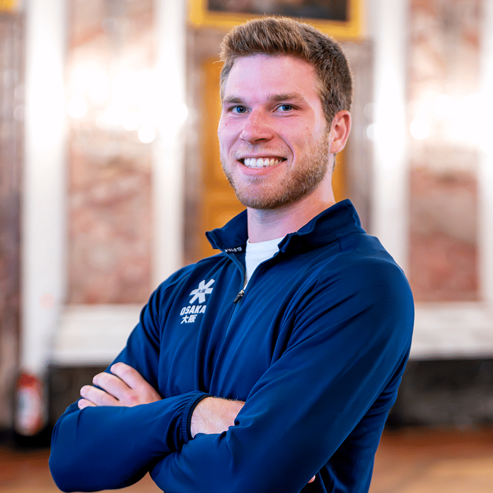 Ein lächelnder Spieler vom MHC in blauer Sportjacke mit verschränkten Armen vor einem prunkvollen Hintergrund für Hockey in Mannheim.