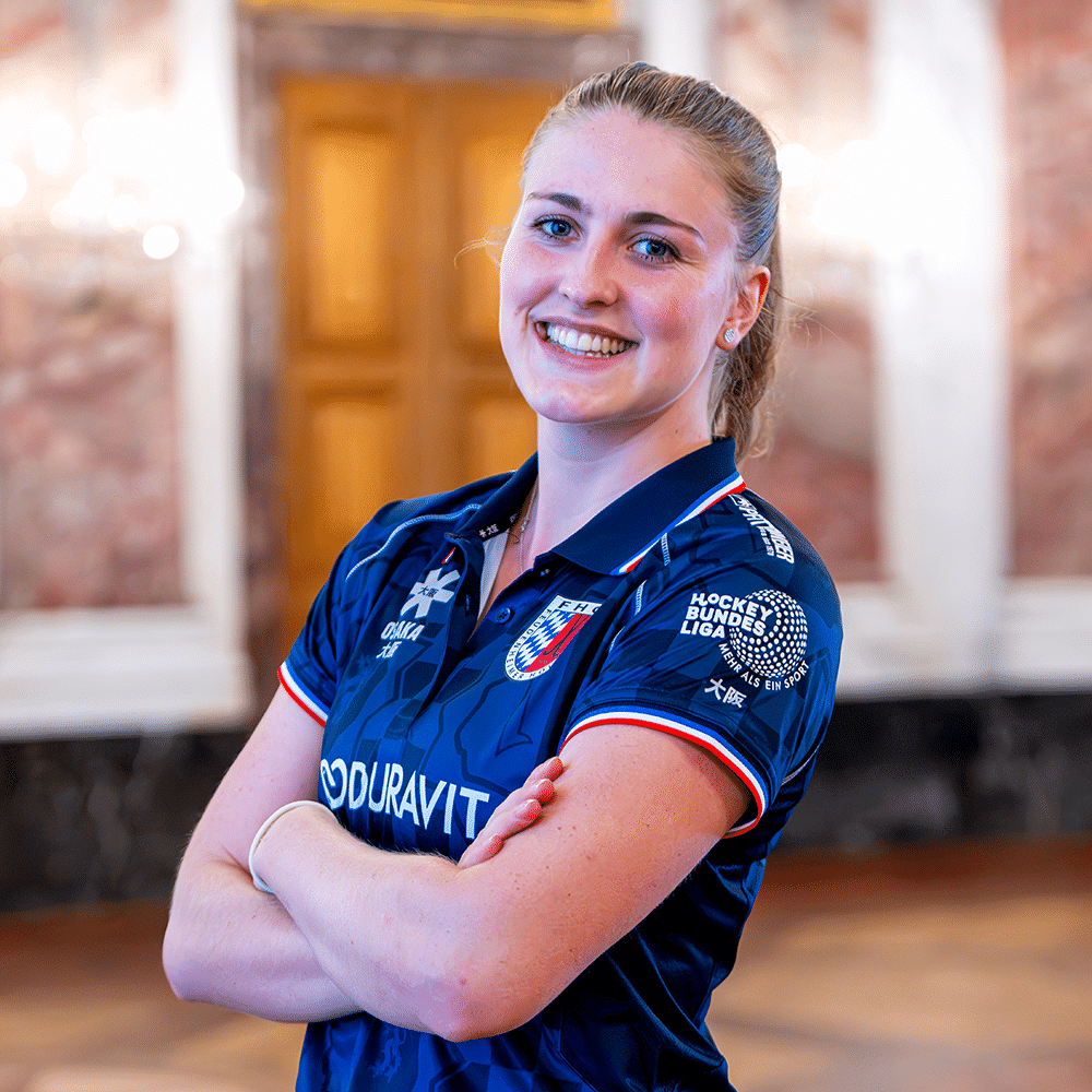 Eine lächelnde Spielerin vom Mannheimer Hockeyclub trägt ein blaues Trikot mit Sponsorenlogos und steht für Hockey in Mannheim vor einer Tür.