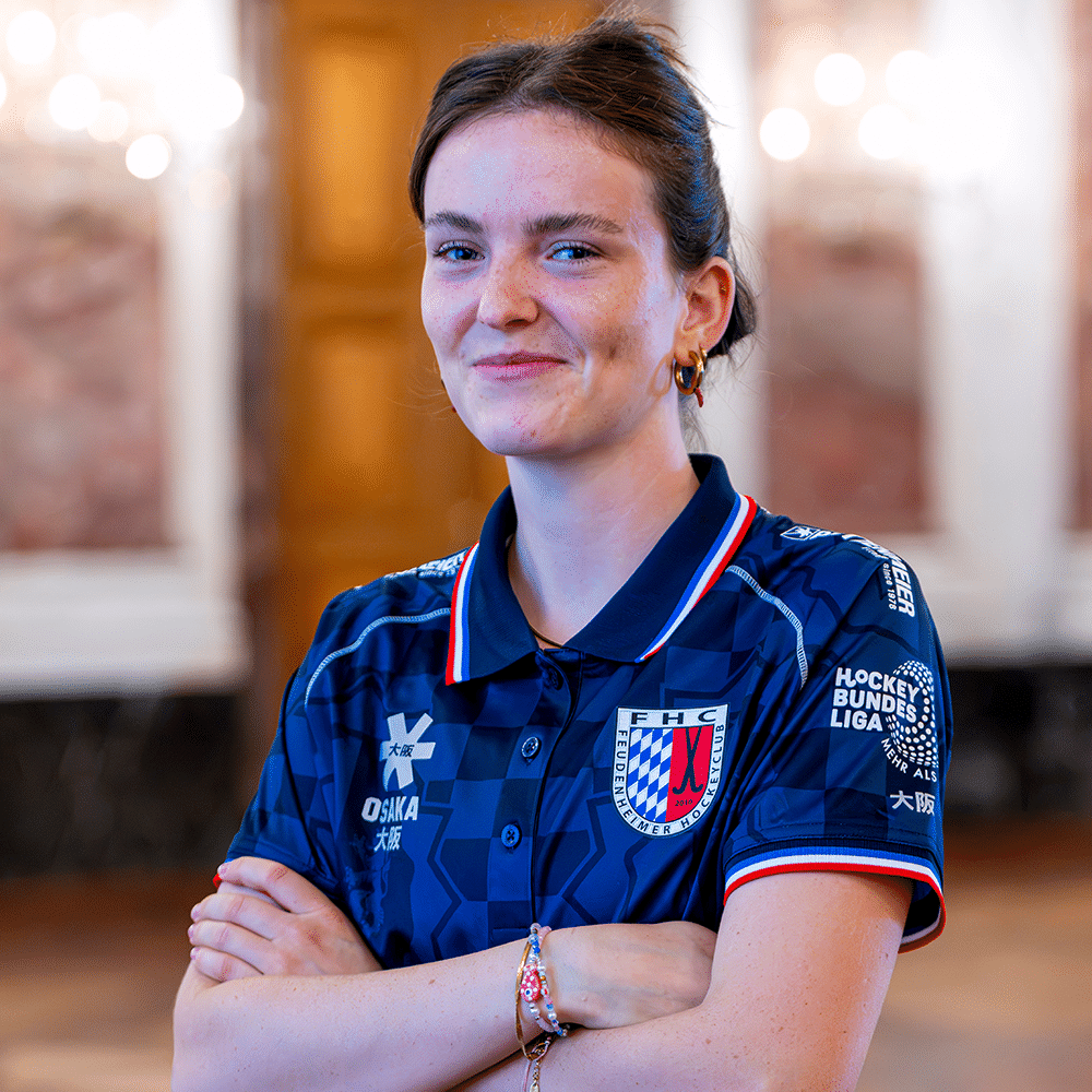 Eine junge Hockeyspielerin des Mannheimer Hockeyclub trägt ein blaues Trikot mit MHC-Wappen und lächelt selbstbewusst in die Kamera.