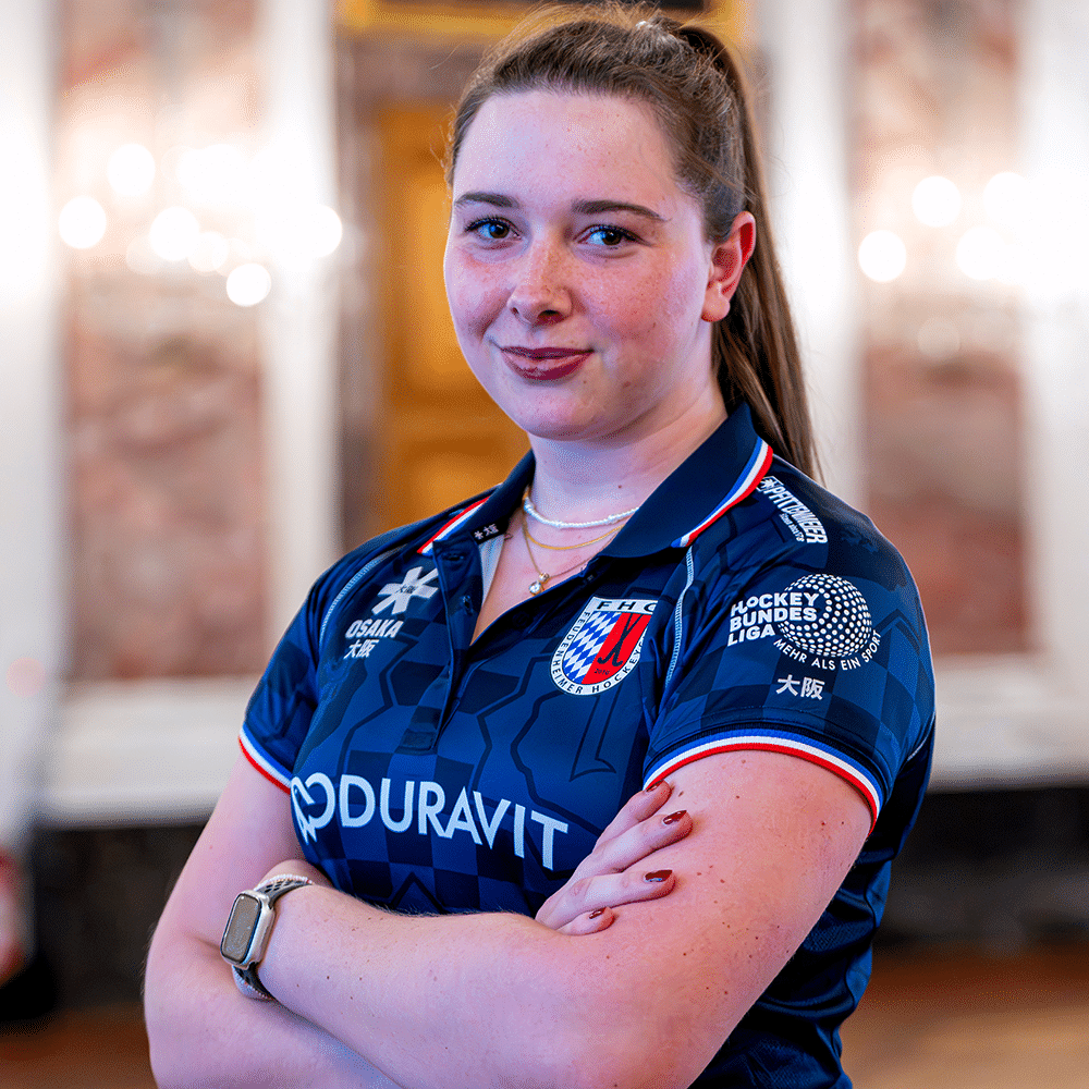 Eine junge Hockeyspielerin vom MHC in ihrem blauen Vereinstrikot mit verschränkten Armen vor einem eleganten, unscharfen Hintergrund.