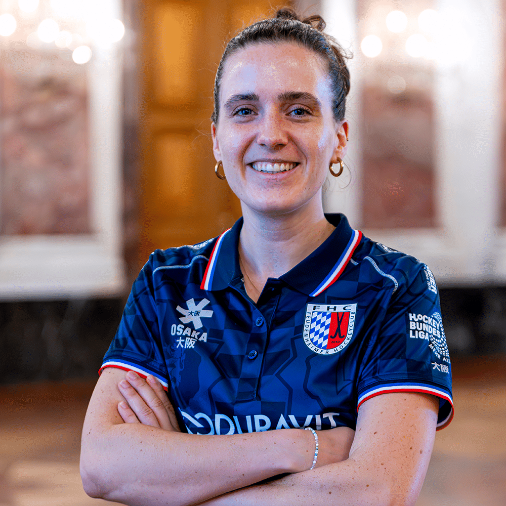 Eine lächelnde Hockeyspielerin in einem blauen Trikot des Feudenheimer Hockeyclubs, die ihre Arme vor der Brust verschränkt hat und in die Kamera blickt.