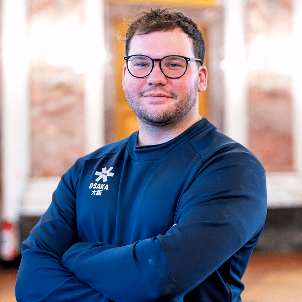 Ein junger Mann mit Brille und dunkelblauem Sportpullover steht mit verschränkten Armen vor einem hellen Hintergrund und lächelt freundlich.