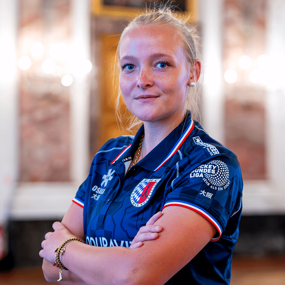 Eine junge blonde Spielerin des MHC trägt das blaue Trikot des Mannheimer Hockeyclub und steht selbstbewusst mit verschränkten Armen vor der Kamera.
