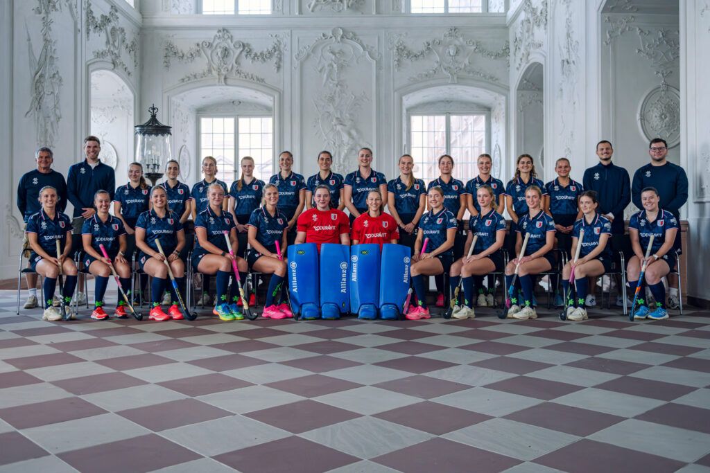 Das Damenteam vom MHC posiert in Hockeytrikots für ein Gruppenfoto in einem prunkvollen Saal. Hockey in Mannheim wird hier professionell repräsentiert.